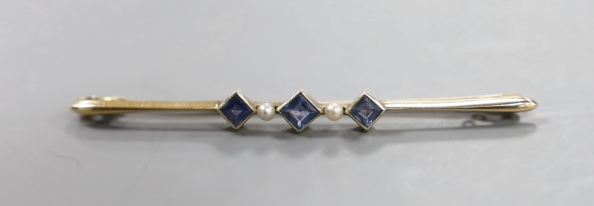 An Edwardian 15ct, three stone sapphire and two stone seed pearl set bar brooch, 57mm, gross 3.4 grams.